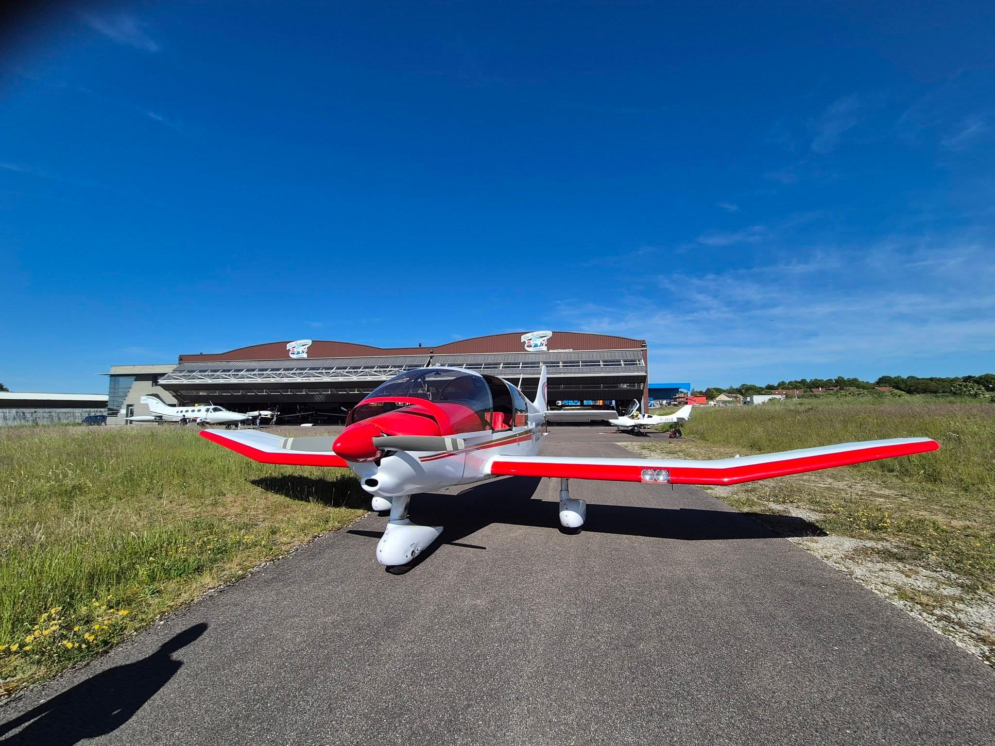 Avion de tourisme rouge stationné sur le taxiway de l'aérodrome de Semur-en-Auxois, devant les hangars, sous un ciel bleu